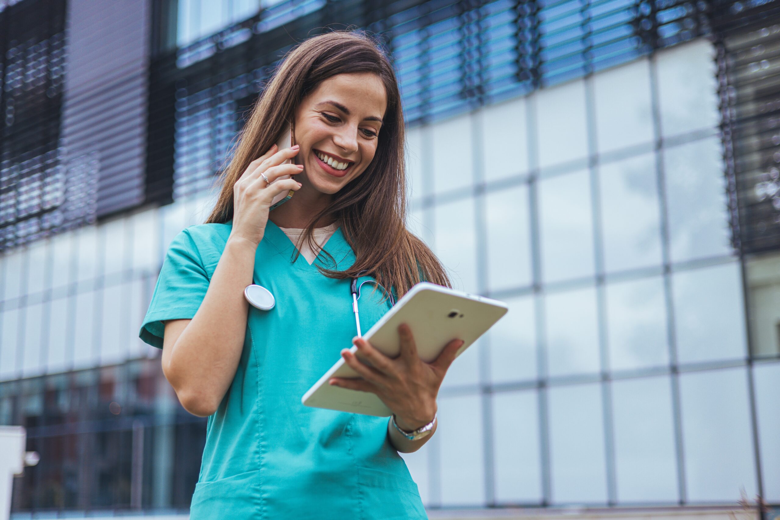 Smiling healthcare worker in scrubs uses a tablet while talking on the phone, set against a modern building backdrop.