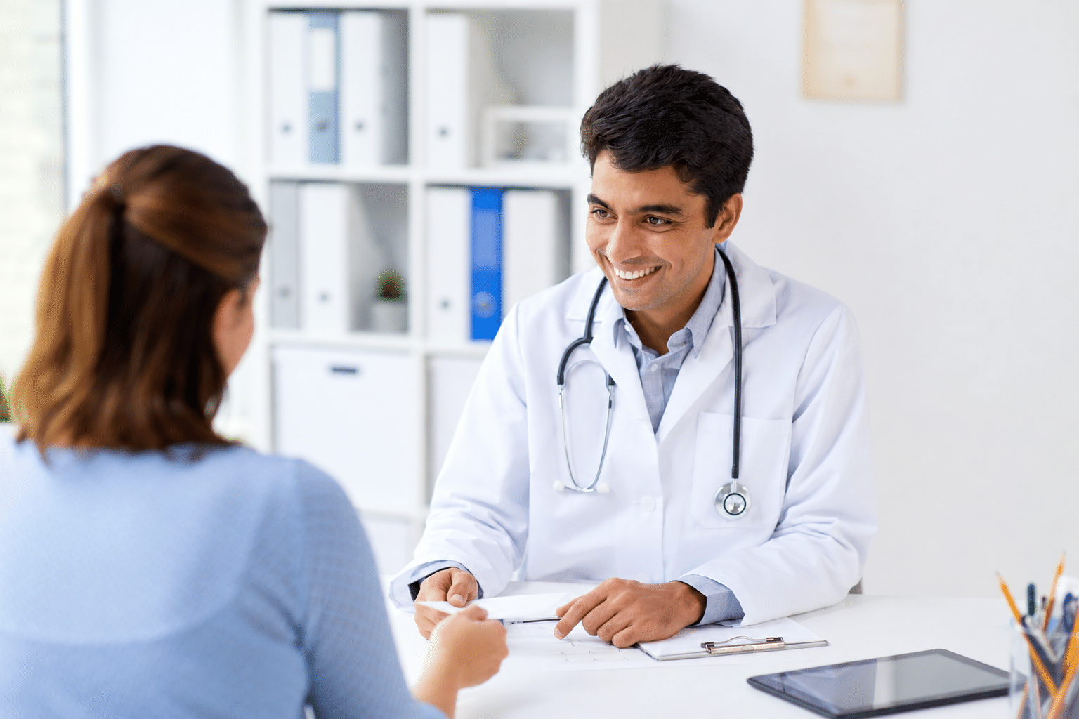A doctor in a white coat smiles at a patient during a consultation. The setting is a bright, organized medical office, emphasizing a welcoming environment.