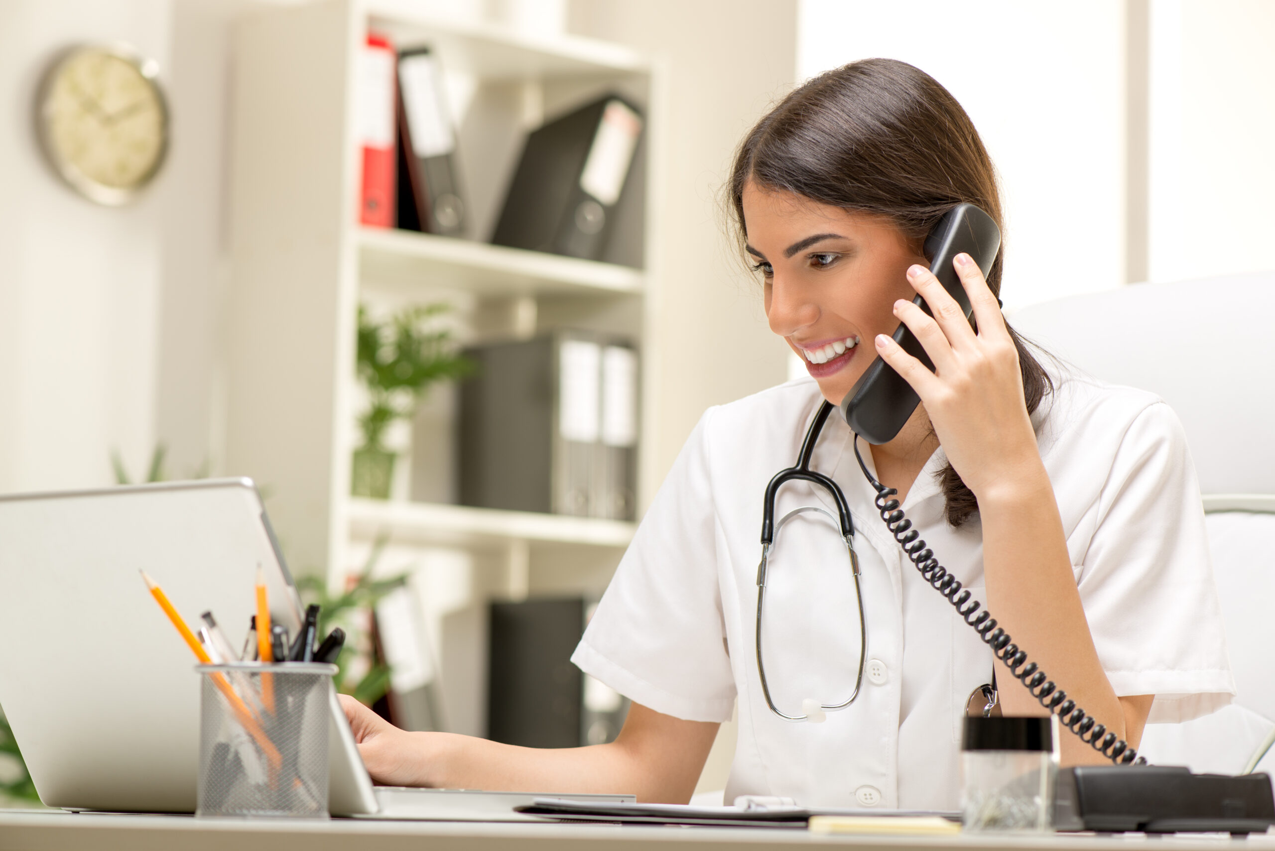 A smiling female doctor on the phone in a modern office, using a laptop. The image conveys a professional and approachable healthcare environment.