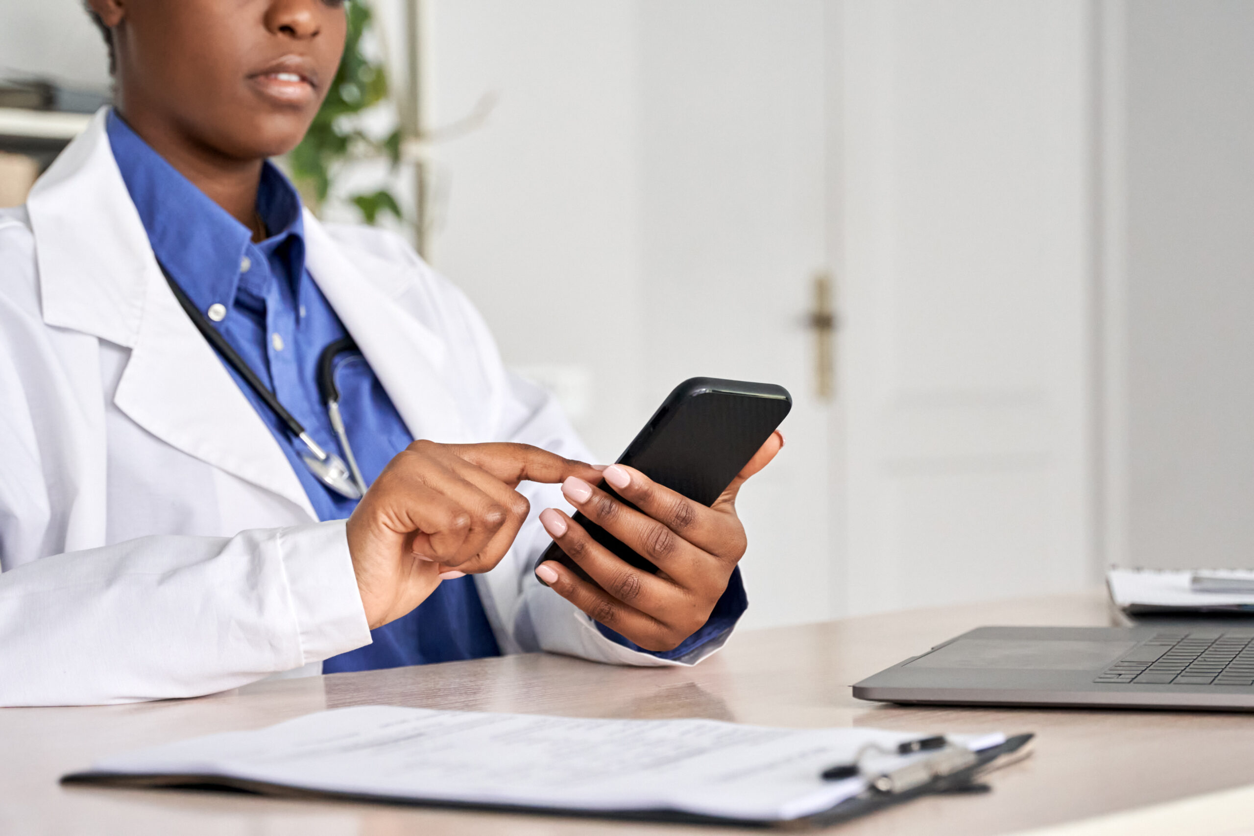 A doctor in a white coat checks a smartphone while seated at a desk with a laptop and documents, highlighting telemedicine or digital health consultations.