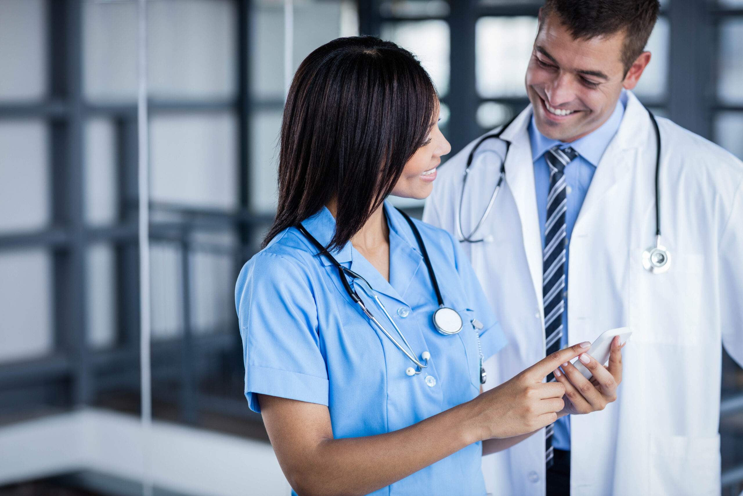 A nurse in scrubs and a doctor in a white coat smile and engage while looking at a smartphone, highlighting teamwork in a healthcare setting.