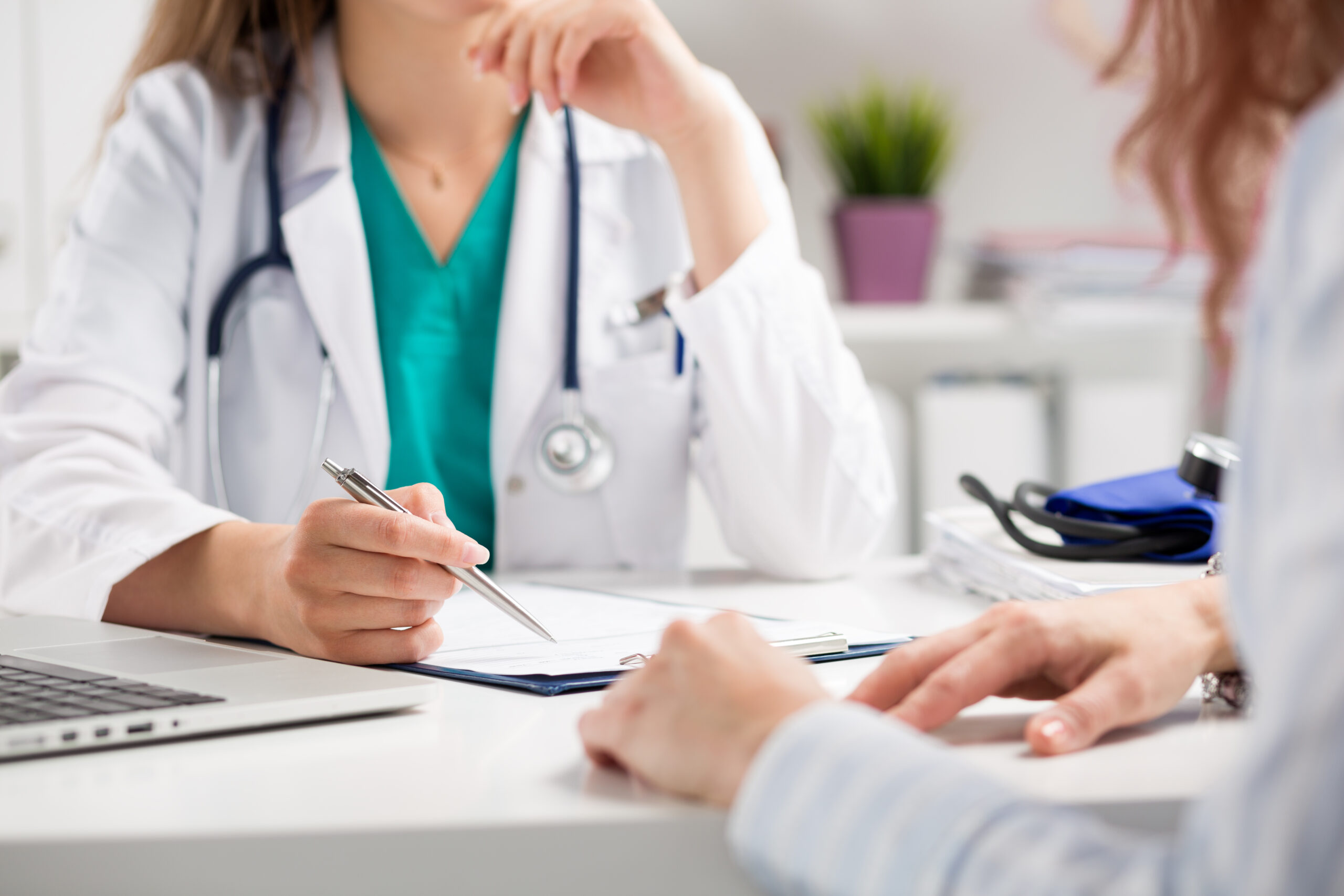 A healthcare professional in a white coat discusses patient information, taking notes with a pen. The setting is a medical office, indicating a consultation.