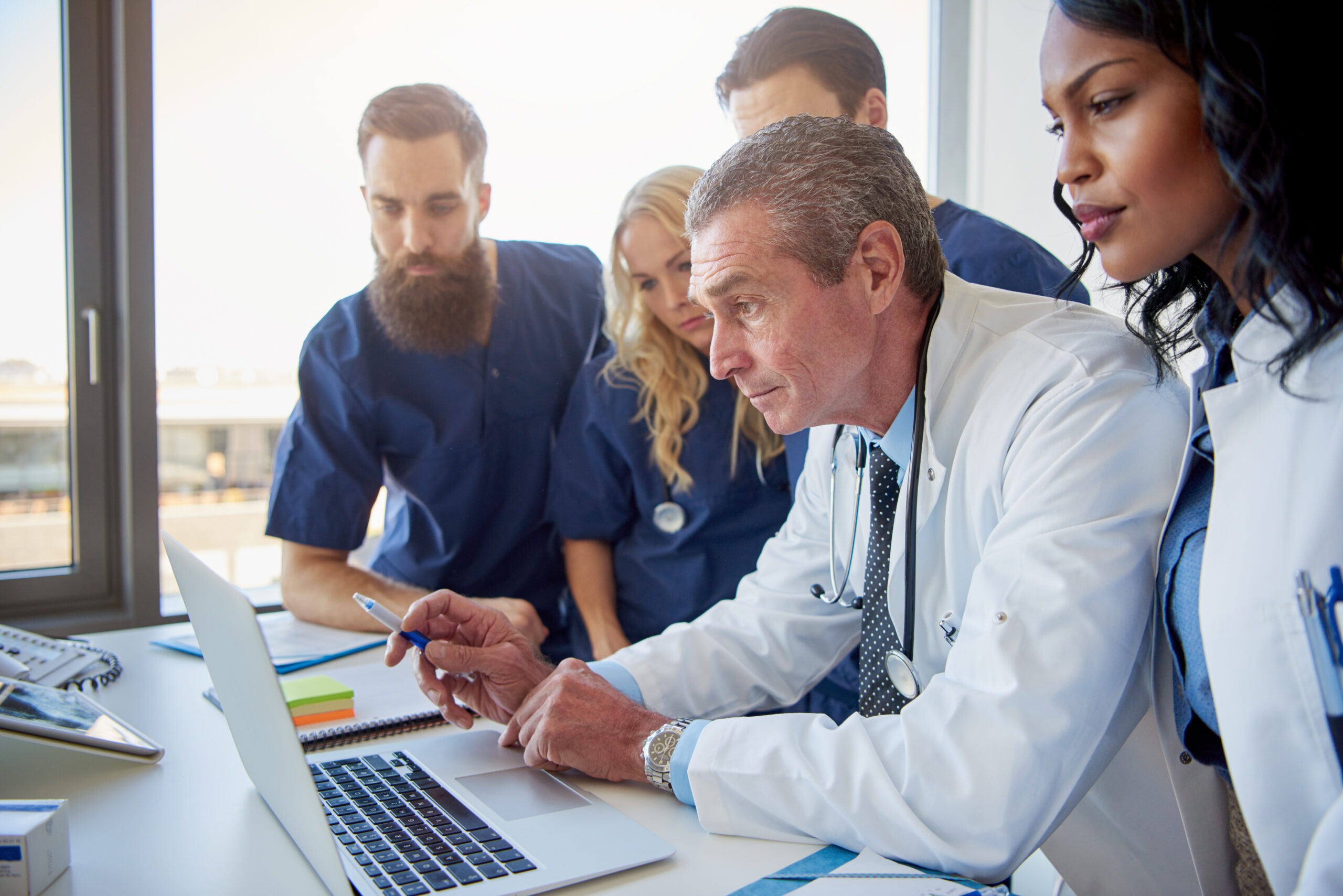 A group of healthcare professionals gathers around a laptop, focused on a discussion led by a doctor in a white coat, emphasizing teamwork in medical decision-making.