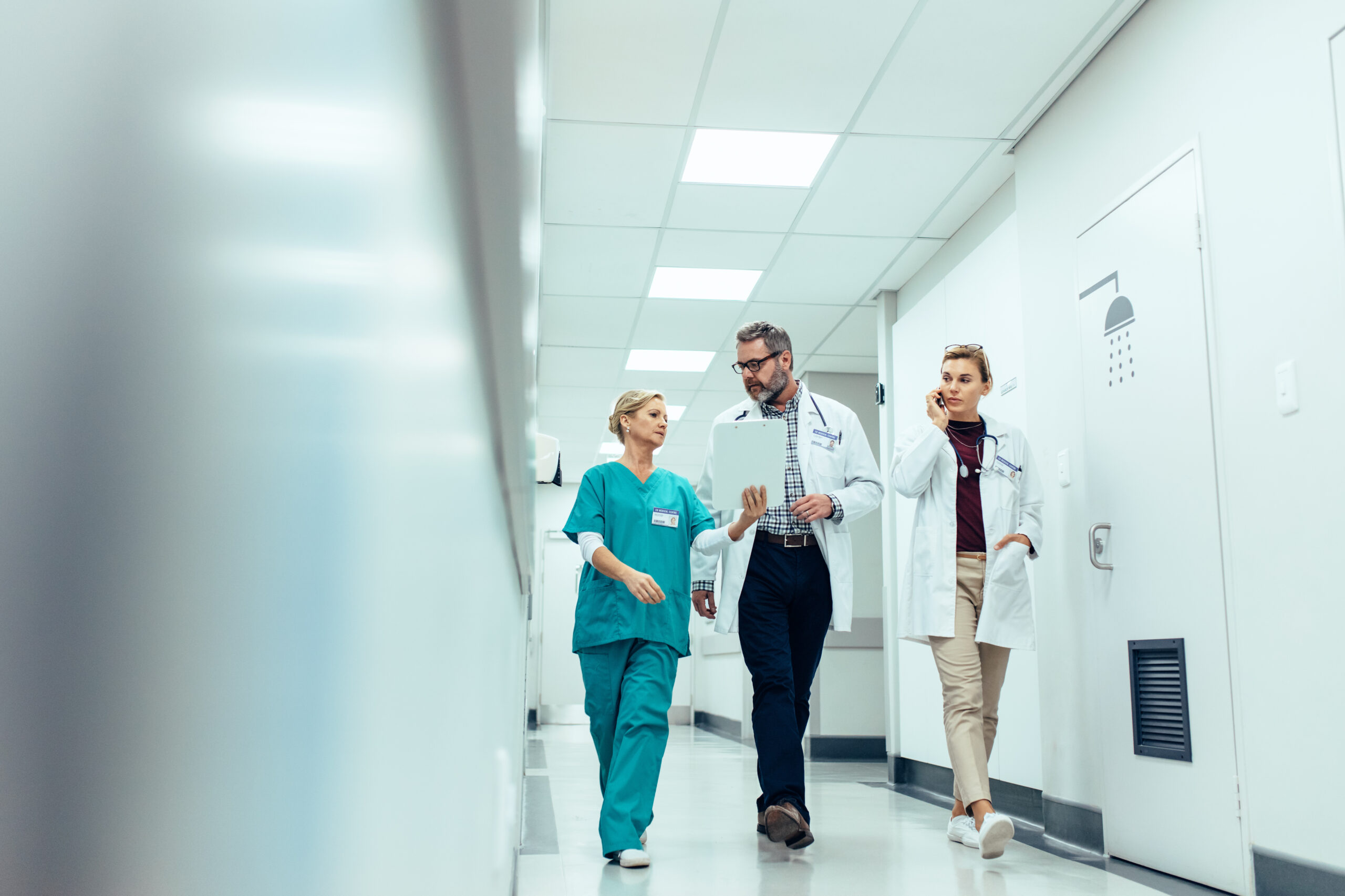 Three medical professionals walk through a hospital hallway. A nurse in teal scrubs discusses patient details with two doctors in white coats.